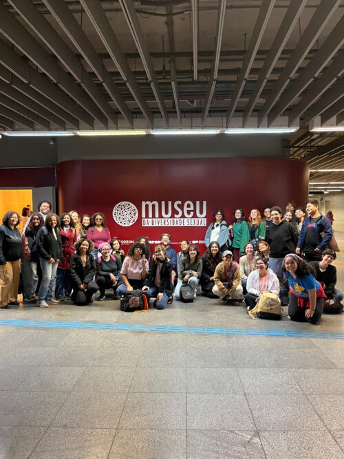 Fotografia que captura um grupo de aproximadamente 30 pessoas. O ambiente é fechado e grande, com piso e teto na cor cinza. Atrás do grupo, há uma grande fachada vermelha com um logotipo e o nome "Museu da Diversidade Sexual" escrito na cor branca. Fim da descrição.