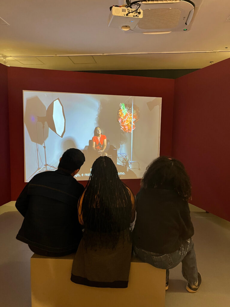 Fotografia tirada em ambiente fechado com iluminação parcial amarela. O teto é branco, o chão é cinza e há uma parede vermelha com um telão em que está sendo projetado um vídeo em que uma pessoa fala dentro de um set de gravações. De frente para o telão e de costas para a câmera, há três pessoas sentadas em um banco assistindo ao vídeo. Fim da descrição.