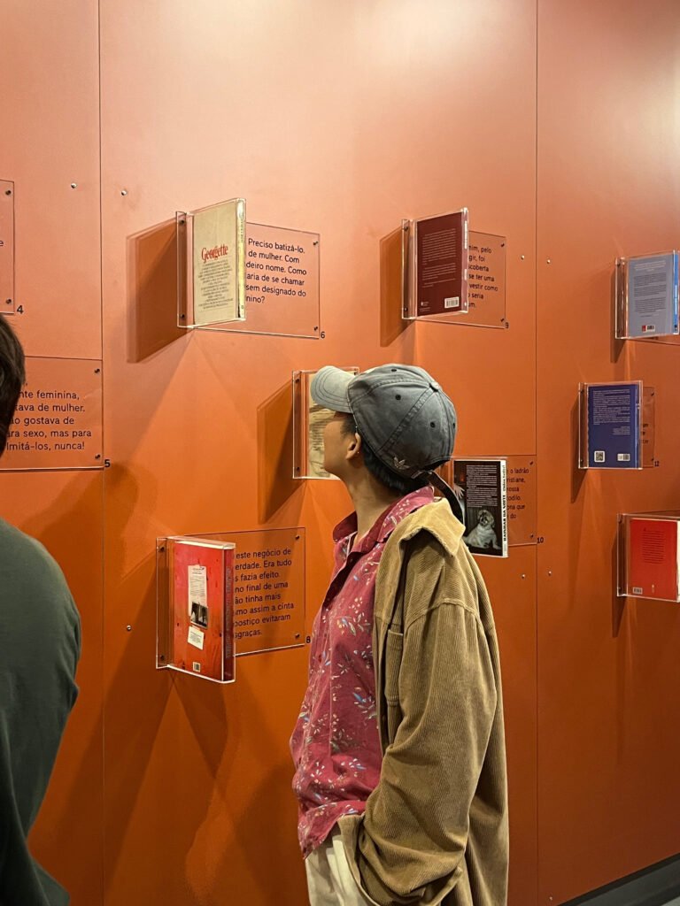 Fotografia tirada em ambiente fechado e bem iluminado. Há uma parede na cor laranja com diversos livros fixados e escritas ao lado. De frente para a parede, há uma pessoa que veste uma camisa vermelha, um casaco marrom e um boné cinza e observa os livros. Fim da descrição.