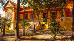 Fotografia que mostra uma construção com arquitetura semelhante á do período colonial do Brasil. A casa é amarela com detalhes vermelhos e há diversas plantas e árvores ao redor. A iluminação é parcial, há luz solar, mas as árvores causam sombra. Fim da descrição.