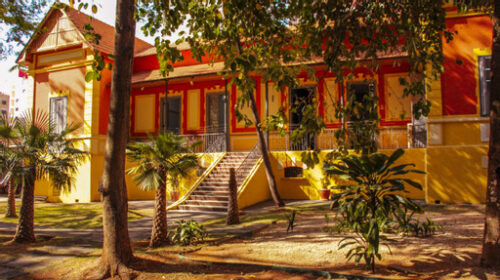 Fotografia que mostra uma construção com arquitetura semelhante á do período colonial do Brasil. A casa é amarela com detalhes vermelhos e há diversas plantas e árvores ao redor. A iluminação é parcial, há luz solar, mas as árvores causam sombra. Fim da descrição.