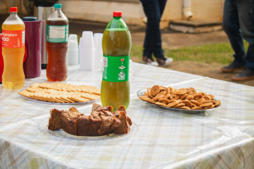 A imagem mostra uma mesa contendo um bolo, biscoitos, refrigetante e copos descartáveis.