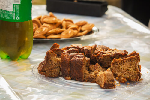 A imagem mostra uma mesa contendo um bolo, biscoitos e refrigetante