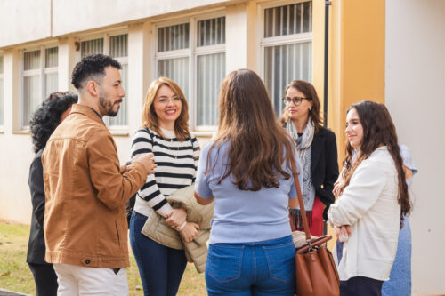 A imagem mostra um grupo de pessoas, 1 homem e 4 mulheres, em circulo conversando. Eles estão em um ambiente externo, de dia.