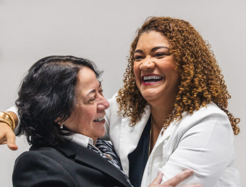 A imagem mostra uma mulher negra, de cabelos cacheados claro sorrindo ao mesmo tempo em que abraça uma mulher branca de cabelos lisos e escuro, também sorrindo. O rosto da primeira é mostrado por inteiro e o da segunda apenas de perfil.