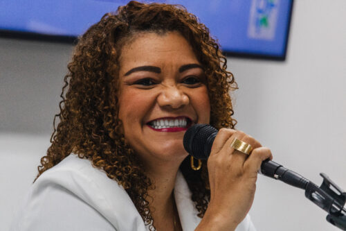 A imagem mostra uma mulher negra de cabelos cacheado castanho claro sorrindo, ela veste um blazer branco e segura um microfone.
