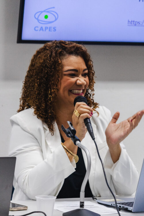 A imagem mostra uma mulher negra de cabelos cacheado castanho claro sorrindo, ela veste um blazer branco e segura um microfone com uma mão