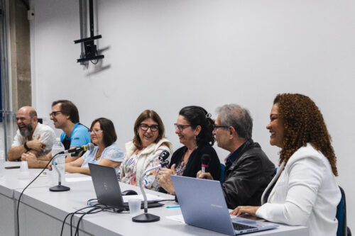 A imagem mostra um grupo de pessoas sentadas de perfil em frente a uma mesa, contendo dois notebooks e microfones. As pessoas sorriem.