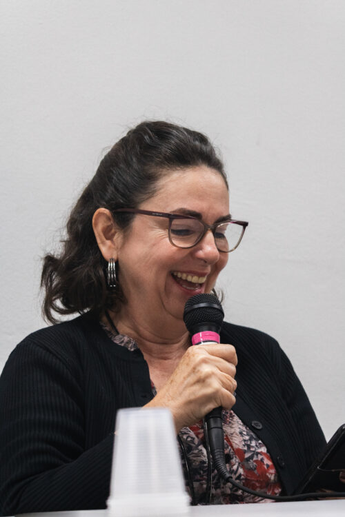 A imagem mostra uma mulher de cabelos pretos usando óculos de grau de armação levemente arredonda na parte inferior e blazer preto. Ela segura um microfone e está sorrindo de cabeça baixa.