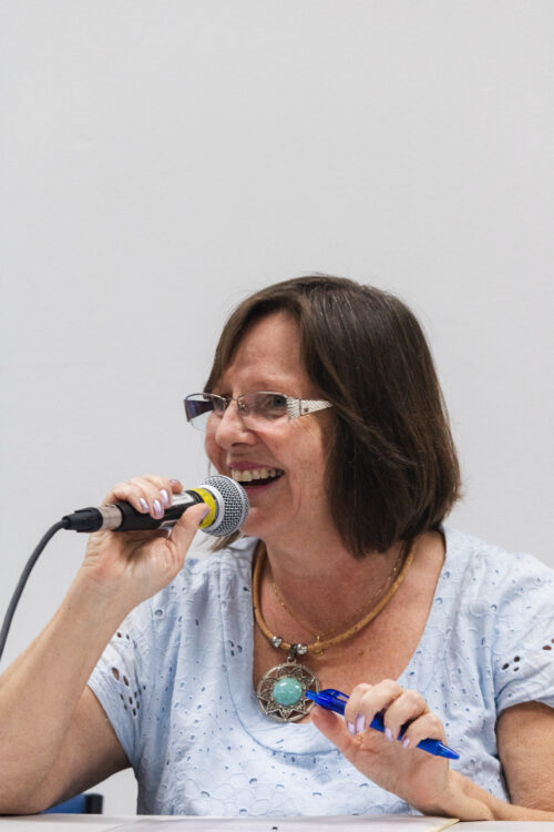 A imamgem mostra uma mulher de cabelos curtos e escuros segurando um microfone com uma mão e uma caneta azul com a outra mão.