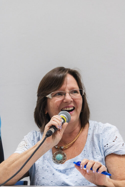 A imamgem mostra uma mulher de cabelos curtos e escuros segurando um microfone com uma mão e uma caneta azul com a outra mão.