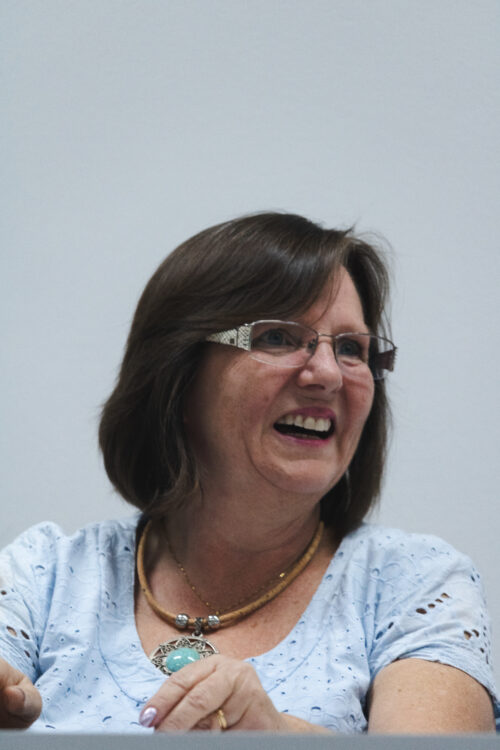 A imagem mostra uma mulher de cabelos escuros e curtos, usando um óculos de grau de armação quadrada e fina, ela está sorrindo.