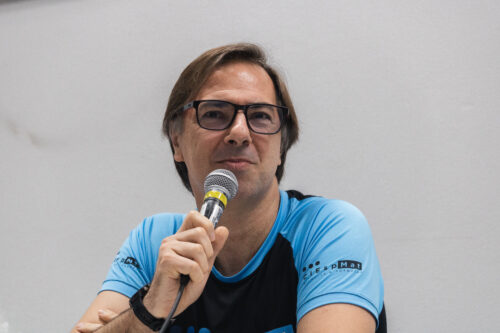 Homem branco de camiseta azul e preta usando oculos de grau quadrado e segurando microfone