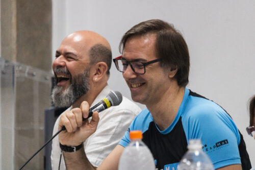 A imagem mostra, em mprimeiro plano, um homem de perfil, usando oculos de grau e camiseta azul. Ao seu lado está um homem barbado de blusa branca. O homem de camiseta azul está sorrindo ao mesmo tempo que segura um microfone o homem barbado está dando risada.