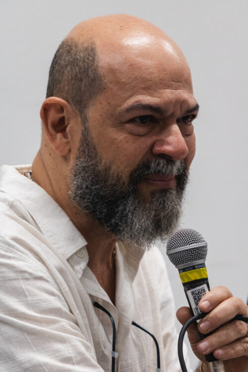 A imagem mostra um homem branco barbado de perifl segurando um microfone.