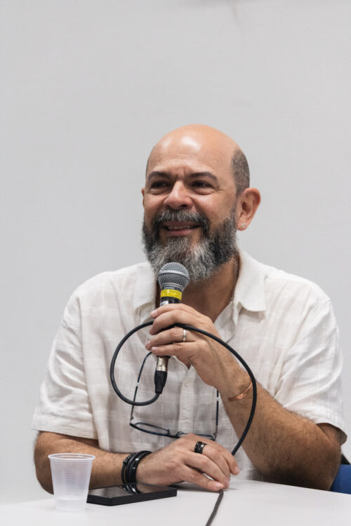 A imagem mostra um homem branco de barba grisalha sorrindo ao mesmo tempo que segura um microfone. Ele está sentado em frente uma mesa, a camera o fotografou de maneira frontal.