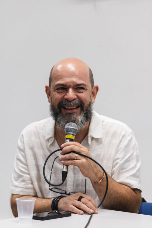 A imagem mostra um homem branco de barba grisalha sorrindo ao mesmo tempo que segura um microfone. Ele está sentado em frente uma mesa, a camera o fotografou de maneira frontal.