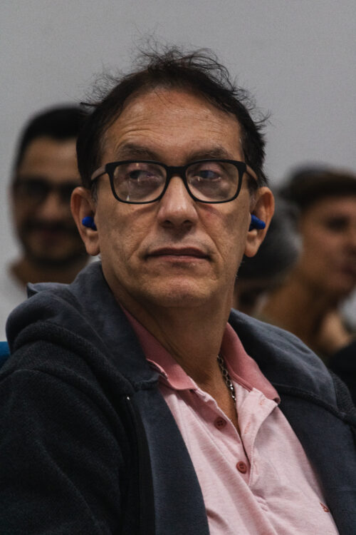 A imagem mostra um homem sentado de frontal, ao fundo há pessoas desfocadas também sentadas.
