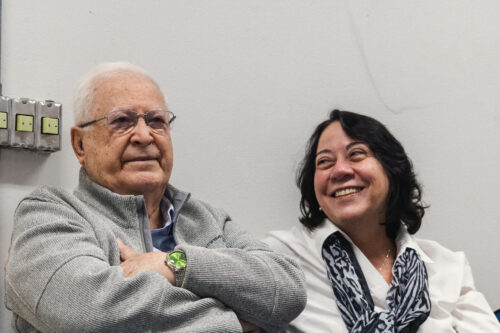 A imagem mostra um homem branco de cabelos brancos com óculos de grau sentado ao lado de uma mulher branca de cabelos curtos e preto. O homem está de braços cruzados, ambos sorriem.