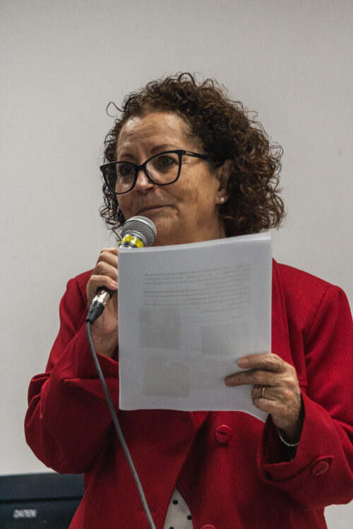 A imagem mostra uma mulher de cabelos cacheados curtos, usando óculos de grau e blazer vermelho. Ela está segurando uma folha sulfite com uma mão e segurando um microfone com a outra.