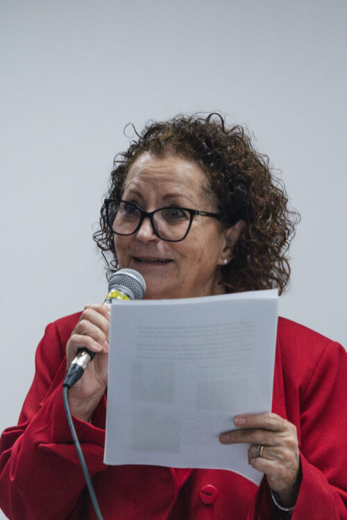 A imagem mostra uma mulher de cabelos cacheados curtos, usando óculos de grau e blazer vermelho. Ela está segurando uma folha sulfite com uma mão e segurando um microfone com a outra.
