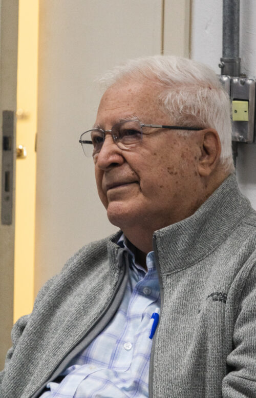 A imagem mostra um homem branco de cabelos brancos usando óculos de grau de armação quadrada e fina, ele veste um casaco cinza e uma blusa azul clara. Ele está sentado de perfil, ao fundo há uma porta entreaberta.