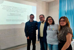Fotografia que captura quatro pessoas em uma sala parciamente iluminada. O grupo está em frente a uma parede com uma projeção com o título "O feminismo negro e a educação do amor no pensamento e na prática de Bell Hooks: movimento de lutas de libertação". Fim da descrição.