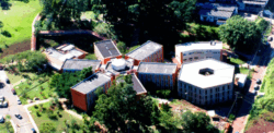 Fotografia aérea que captura o prédio da Faculdade de Educação da Unicamp durante o dia. O prédio tem formato de asterisco e há dois anexos à sua direita, um em formato de hexágono e o outro em retângulo. Há grama e muitas árvores em volta dos prédios. Fim da descrição.