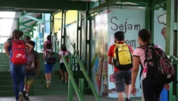 Fotografia que captura alunos entrando em escola. As paredes têm grafites e o ambiente tem iluminação parcial. Fim da descrição.
