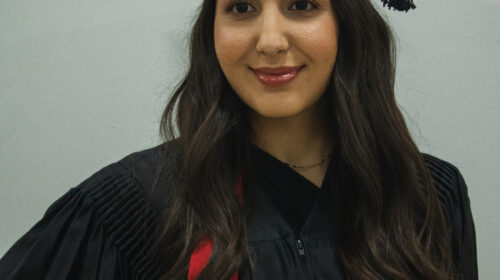 Uma jovem de pele clara, com cabelos castanhos longos e ondulados, está sorrindo levemente e olhando para a esquerda. Ela veste uma beca preta de formatura, com um capelo na cabeça e uma faixa vermelha sobre os ombros. A beca tem pregas nas mangas e um zíper frontal. A faixa vermelha se destaca sobre o preto da vestimenta. O fundo da imagem é uma parede branca, e a iluminação suave foca na pessoa, que está com a mão direita na cintura.