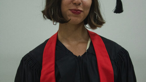 Formanda usando beca, capelo e faixa vermelha. Ela é branca, de cabelos loiros e curtos e sorri para a foto discretamente.
