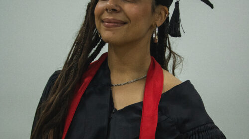 Formanda usando beca, capelo e faixa vermelha. Ela é negra de pele clara, de cabelos trançados e longos e sorri para a foto discretamente