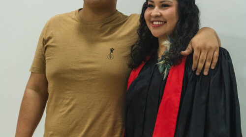 ois jovens pardos estão lado a lado em um fundo branco, posando para uma foto. O rapaz, à esquerda, tem cabelo curto e escuro, e veste uma camiseta mostarda e calça jeans clara, com o braço esquerdo sobre o ombro da garota. A jovem, à direita, tem cabelos longos e ondulados, e veste uma beca de formatura preta com uma faixa vermelha, além de um capelo com borla. Ela sorri, e uma pequena flor branca e verde está presa à sua beca. Ambos olham para a esquerda, fora da câmera, com a iluminação suave vindo da parte superior do ambiente.
