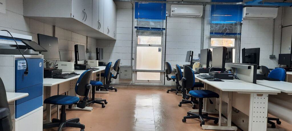 Fotografia que mostra laboratório de informática. O ambiente é bem iluminado, tem paredes claras e piso marrom. Há diversas estações de trabalho com computadores, teclados e mouses, além de cadeiras com estofados azuis escuro. Acima dos espaços de trabalho, anexados à parede, há armários. Também fixado na parede, há um aparelho de ar condicionado. Fim da descrição.