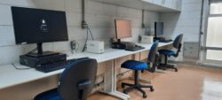 Fotografia que mostra duas três estações de trabalho em um laboratório de informática. O ambiente é bem iluminado, as paredes são claras e o piso é marrom. Há uma mesa comprida que comporta as três estações. Cada estação tem um computador, um teclado, um mouse e uma cadeira com estofado azul escuro. Os computadores são grandes e modernos. Acima das estações, anexados na parede, há armários. Fim da descrição.