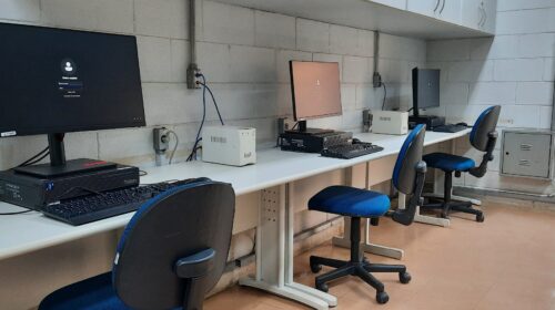 Fotografia que mostra duas três estações de trabalho em um laboratório de informática. O ambiente é bem iluminado, as paredes são claras e o piso é marrom. Há uma mesa comprida que comporta as três estações. Cada estação tem um computador, um teclado, um mouse e uma cadeira com estofado azul escuro. Os computadores são grandes e modernos. Acima das estações, anexados na parede, há armários. Fim da descrição.