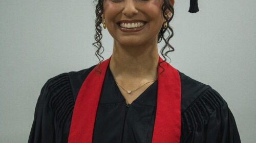Formanada usando beca, capelo e faixa veremelha no pescoço. Ela está posicionada de maneira frontal para a camera, sorrindo. Duas mechas cacheadas escapam de seus cabelos presos.