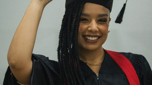 Uma jovem negra de pele clara com cabelos em tranças longas e escuras está sorrindo de forma alegre, segurando a borda do capelo de formatura que está na sua cabeça. Ela veste uma beca preta com uma faixa vermelha sobre os ombros e possui um colar dourado no pescoço. No seu rosto, há um piercing no nariz e um leve brilho nos lábios. O fundo da imagem é uma parede lisa em tom cinza-claro.