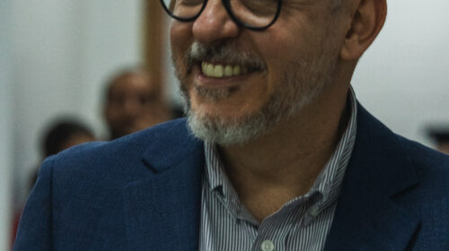 Homem branco de cabelos e cavanhaque grisalho. Ele sorri com os olhos entreabertos. Usa óculos pretos, terno azul e camisa azul.