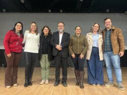 Fotografia que mostra um grupo de sete pessoas, cinco mulheres e dois homens, todos sorrindo, alguns deles vestem roupas formais, outros vestem roupas causais. O ambiente é fechado e se assemelha a um auditório; o grupo está acima de um palco, o piso do palco é de madeira; o ambiente é bem iluminado. Fim da descrição.