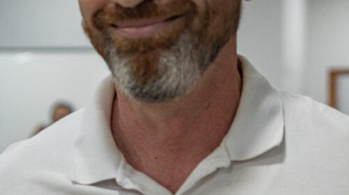 Homem branco de cabeços e barba grisalha. Ele usa uma camiseta polo branca.