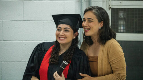 Duas mulheres sorriem olhando para algo extraquadro. O fundo mostra uma parede branca, a iluminação é artificial branca, indicando ambiente interno. A mulher da direita, branca de vestes amarronzadas, segura delicamente o braço da outra. A outra, posicionada levemente a frente da primeira, usa beca e capelo, sorrindo.