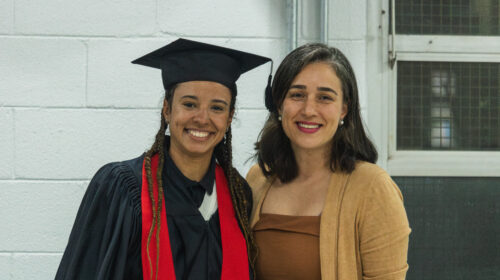 Duas mulheres sorriem para a foto. O fundo é uma parede branca e a iluminação é artificial branca. Uma das mulheres, negra, veste uma beca e capelo a outra, mulher branca, veste roupas em tons amarronzados.