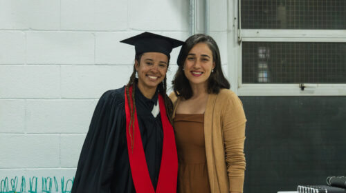 Duas mulheres sorriem para a foto. O fundo é uma parede branca e a iluminação é artificial branca. Uma das mulheres, negra, veste uma beca e capelo a outra, mulher branca, veste roupas em tons amarronzados.