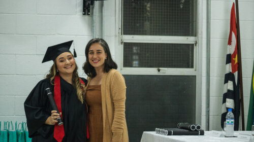 Duas mulheres sorriem para a foto. Estão posicionadas de frente, ocupando apenas 1/3 da imagem. O restante da imagem é ocupado por um cenário interno, composto por uma mesa branca, janela e parede branca. Uma mulher, branca de cabelos escuros veste roupa em tons amarronzados a outra, mulher loira usa beca e capelo ao mesmo tempo em que segura um canudo.