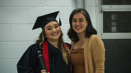 Duas mulheres sorriem para a foto. A iluminação é artificial branca. Ao fundo, é possível notar paredes brancas. A primeira mulher, loira, veste beca e capelo ao mesmo tempo em que segura um canudo. A outra, mulher branca, de cabelos escuros pretos veste roupas amarronzadas ao mesmo tempo em que abraça a formanda.