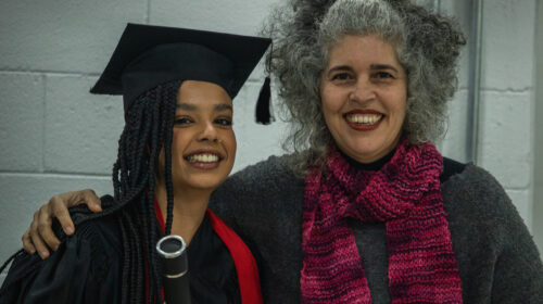 Duas mulheres sorriem para a foto. Uma, mulher negra de cabelos trançados longos, veste uma beca e capelo enquanto segura um canudo. A outra mulher branca de cabelos grisalhos veste uma blusa cinza e cachecol rosa ao mesmo tempo em que abraça a outra.