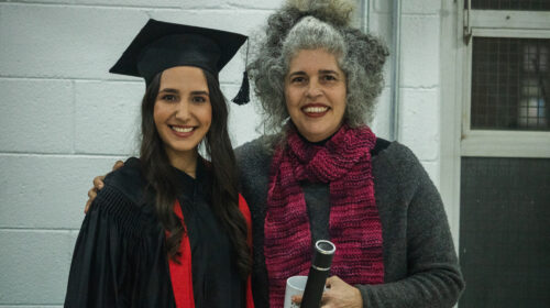 uas mulheres sorriem para a foto. Uma, mulher negra de cabelos trançados longos, veste uma beca e capelo enquanto segura um canudo. A outra mulher branca de cabelos grisalhos veste uma blusa cinza e cachecol rosa ao mesmo tempo em que abraça a outra.
