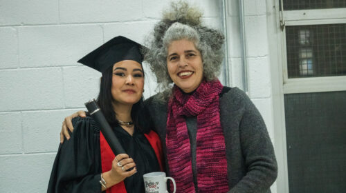 uas mulheres sorriem para a foto. Uma, mulher negra de cabelos trançados longos, veste uma beca e capelo enquanto segura um canudo. A outra mulher branca de cabelos grisalhos veste uma blusa cinza e cachecol rosa ao mesmo tempo em que abraça a outra.