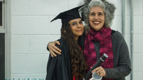 uas mulheres sorriem para a foto. Uma, mulher negra de cabelos trançados longos, veste uma beca e capelo enquanto segura um canudo. A outra mulher branca de cabelos grisalhos veste uma blusa cinza e cachecol rosa ao mesmo tempo em que abraça a outra.